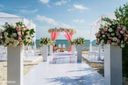 Outdoor beach wedding setup with white chairs, flower arrangements, and a pink-draped altar facing the ocean.
