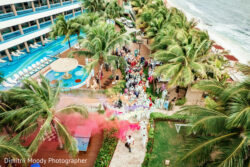 A wedding party gathers outdoors by the beach as pink smoke fills the air, surrounded by palm trees and a resort.