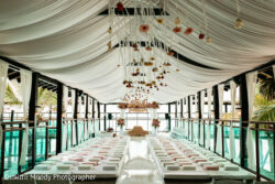 Indoor wedding aisle with white draped ceiling, hanging flowers, and rows of cushions on the floor facing a floral altar.