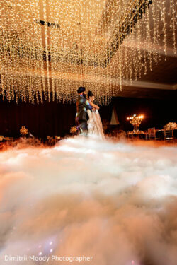A couple dances surrounded by low-lying fog and string lights in a decorated indoor venue.
