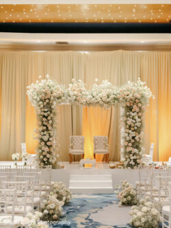 Rushika & Neil, JW Marriot Cancun Mexico, Wedding Ceremony Indoor wedding stage with two chairs under a floral arch, surrounded by white flowers and rows of chairs.