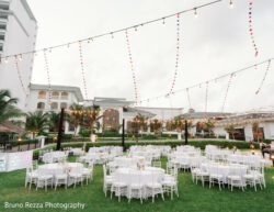 Rushika & Neil, JW Marriot Cancun Mexico, Welcome Party Outdoor event setup with round tables and white chairs on grass, string lights and colorful garlands overhead.