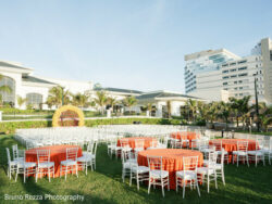 Rushika & Neil, JW Marriot Cancun Mexico, Hauldi Outdoor event setup with round tables covered in orange cloths and white chairs on a lawn, with buildings in the background.