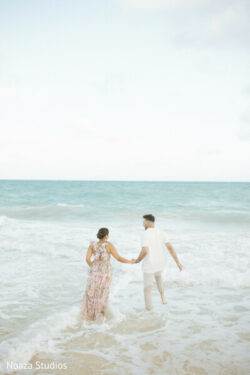 A couple holding hands walks into the ocean waves at a beach under a cloudy sky.