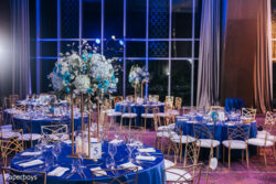 Elegant banquet hall with round tables covered in blue cloths, gold chairs, and tall floral centerpieces.