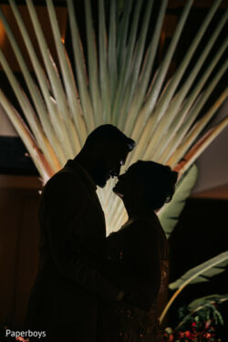A silhouetted couple stands close together in front of a large, fanned palm leaf backdrop.