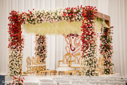 A wedding mandap decorated with red, white, and pink flowers, featuring gold chairs and a white backdrop.