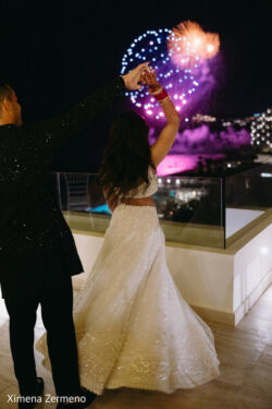 A couple in formal attire dances on a balcony at night, watching fireworks over a distant city and waterfront.