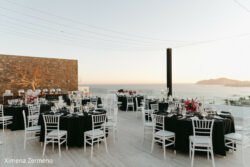 Outdoor event space with round tables covered in black cloths, white chairs, and ocean view at sunset.