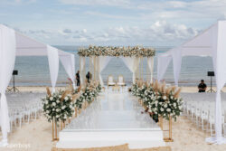 An outdoor beach wedding setup with white chairs, floral arrangements, and a decorated stage facing the ocean.