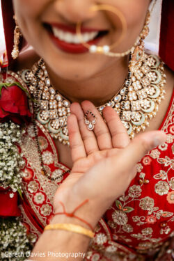 A bride in red attire smiles as a hand holds a mangalsutra near her ornate necklace and nose ring.