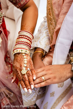 Close-up of a bride and groom's hands at a wedding, showing henna, bangles, and wedding rings.