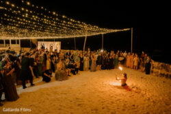 A fire dancer performs on sand at night for a large crowd under string lights at an outdoor event.