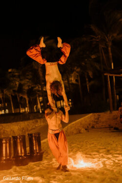 Two male performers in orange pants do a hand-balancing act on a sandy beach at night near a fire.