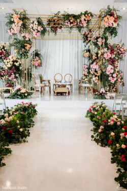 A wedding mandap decorated with pink and white flowers, gold framework, and two chairs on a raised white platform.