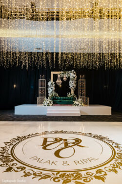Wedding stage with floral decor and a green sofa, under hanging lights; "Palak & Ripal" written on the floor.