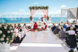 A Sikh wedding ceremony takes place outdoors by the ocean, with guests seated around the mandap under a clear blue sky.