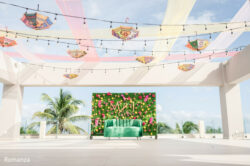 A decorated outdoor stage with a green couch, flower wall, and hanging umbrellas under string lights.