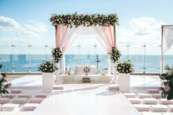 Outdoor wedding mandap with pink and white drapes, floral arrangements, and a view of the ocean in the background.