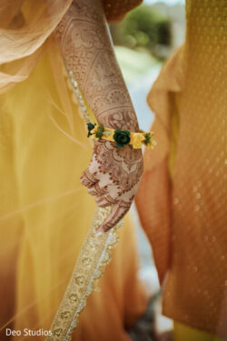 A hand with intricate henna designs and a yellow floral bracelet, wearing a yellow outfit with gold trim.