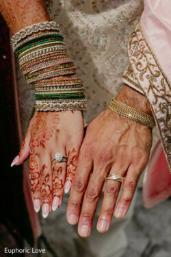 A close-up of a couple’s hands showing wedding rings, bangles, and henna designs, dressed in traditional attire.